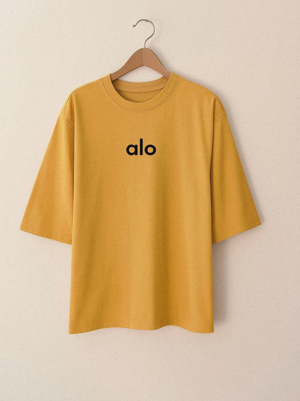 تیشرت زنانه با رنگ گرم خردلی و طراحی مینیمال، دارای نوشته alo به صورت ظریف روی سینه و آستینهایی تا سطح آرنج، پسزمینهای ساده که حس تابستانی و مدرن را القا میکند.